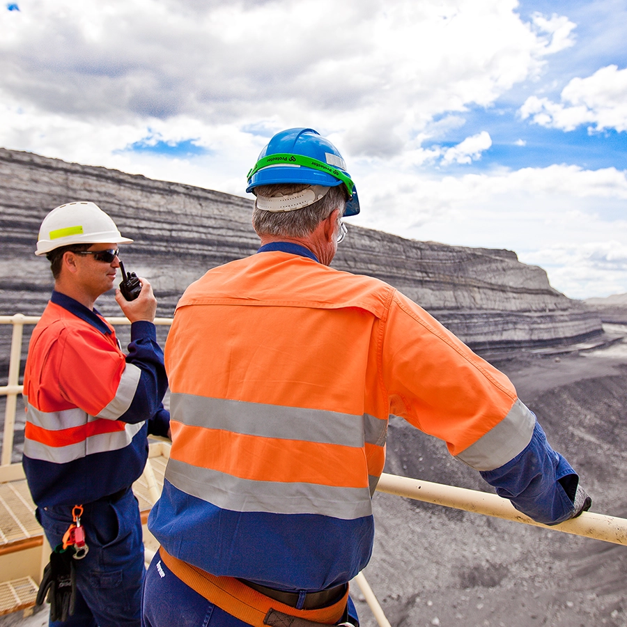 Two workers in safety gear overlooking an open-pit mine, one holding a two-way radio.