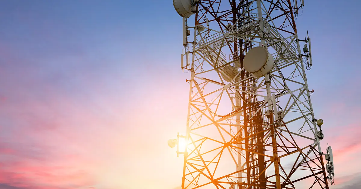 Communication tower with antennas and satellite dishes silhouetted against a colourful sunrise or sunset sky.