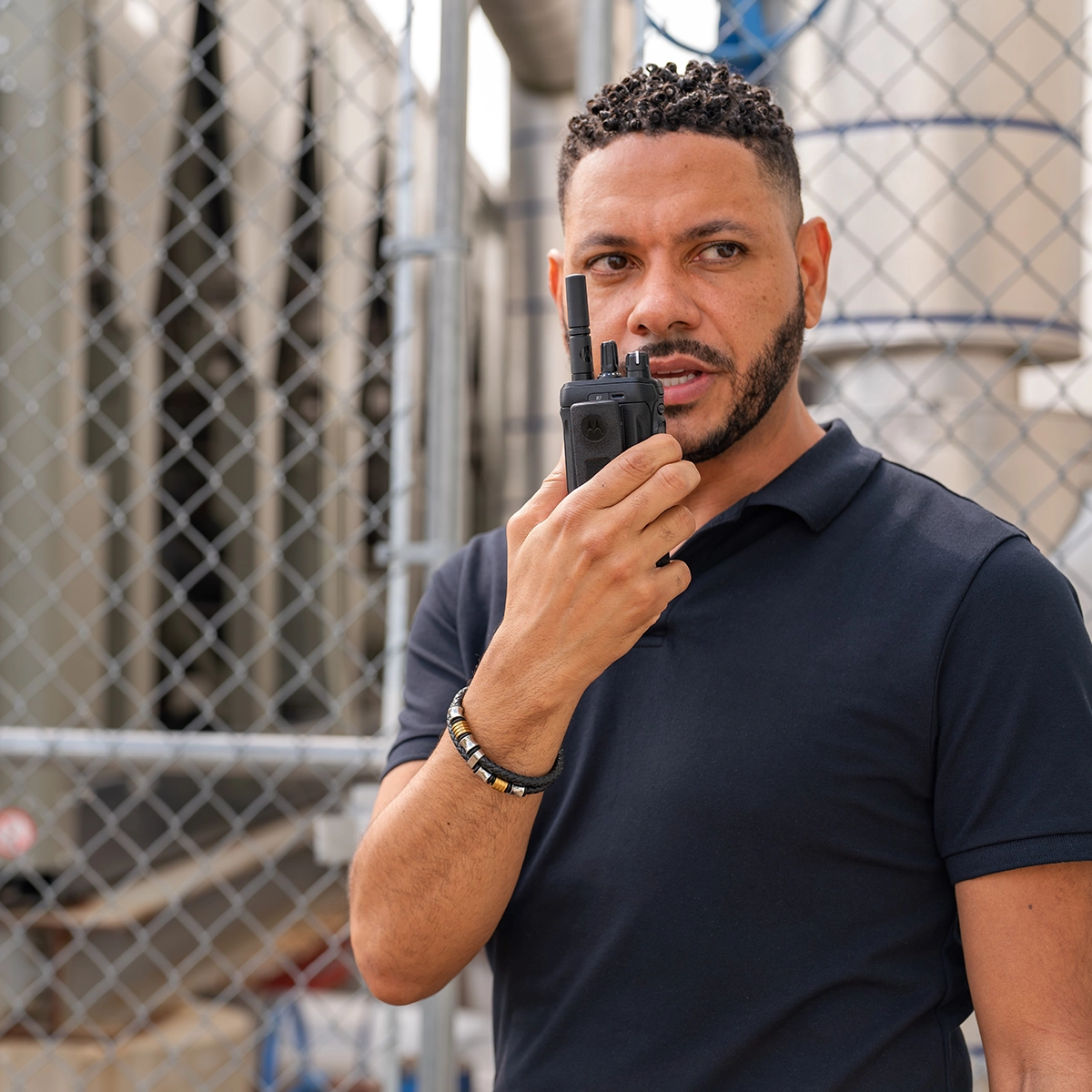 Individual holding a DMR two-way radio near industrial equipment behind a chain-link fence.
