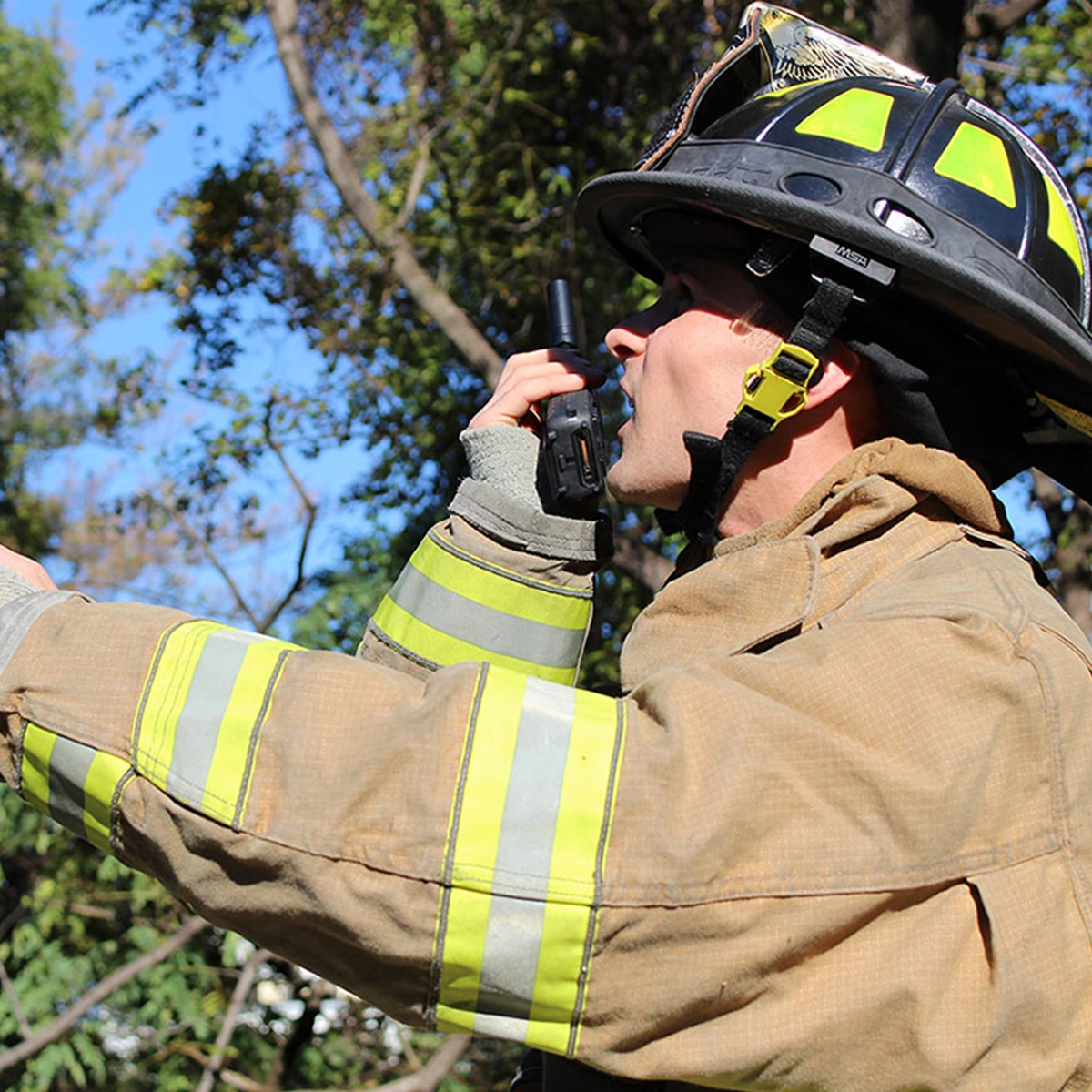 Firefighter in full protective gear using a push-to-talk radio in an outdoor setting with trees in the background.