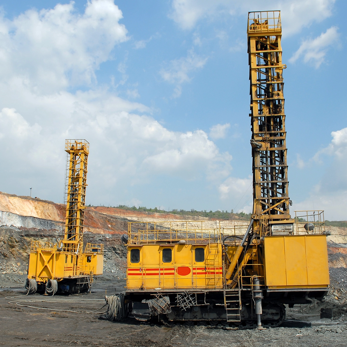 Heavy-duty drilling rigs in a rocky open-pit mining site with exposed earth layers and industrial equipment.