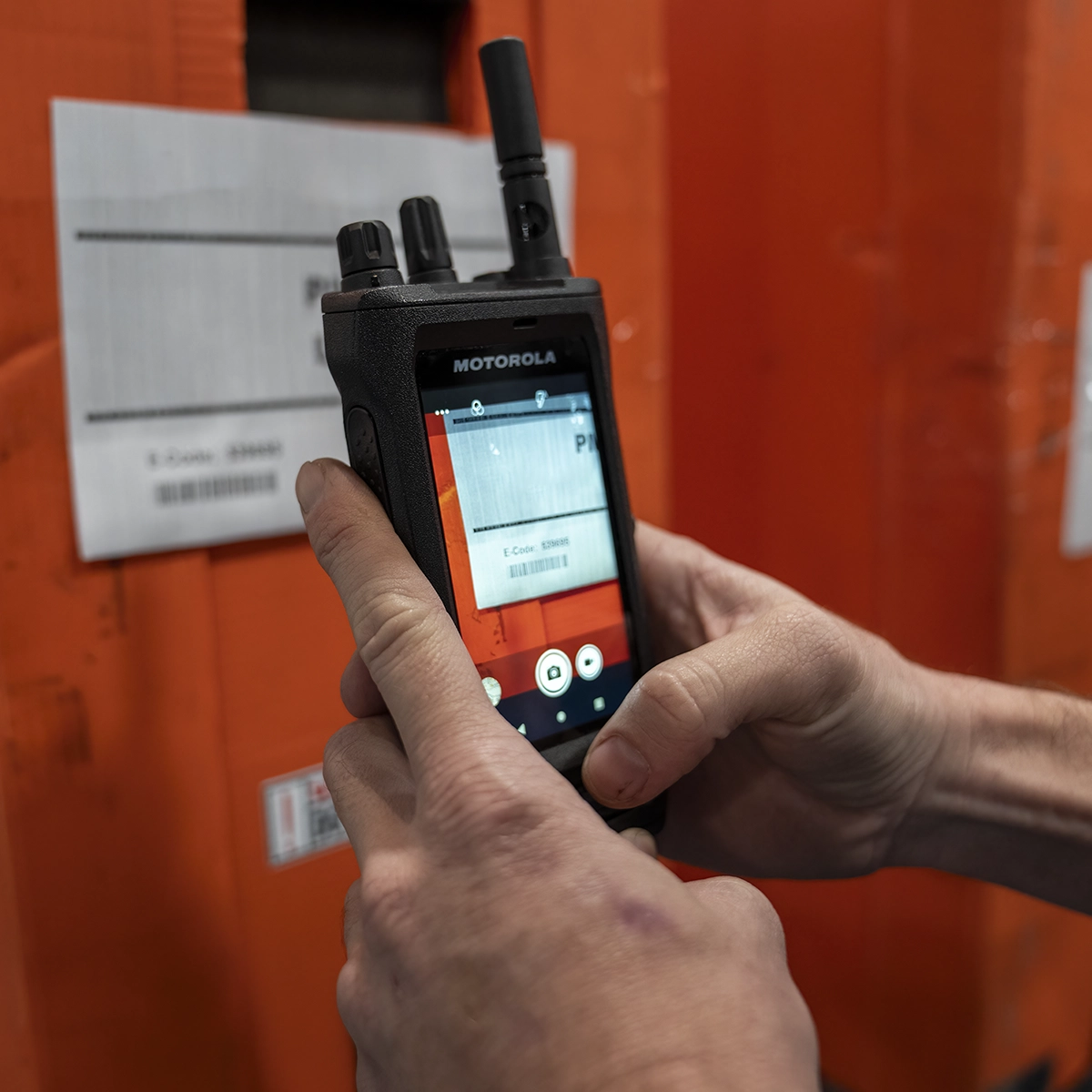 Person using a Motorola MOTOTRBO Ion Smart Radio to scan a barcode label on an orange background.
