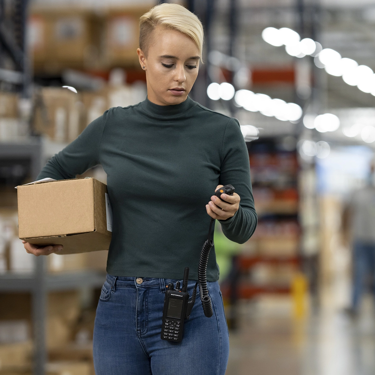 Warehouse worker holding a box and using a two-way radio in an industrial storage area.