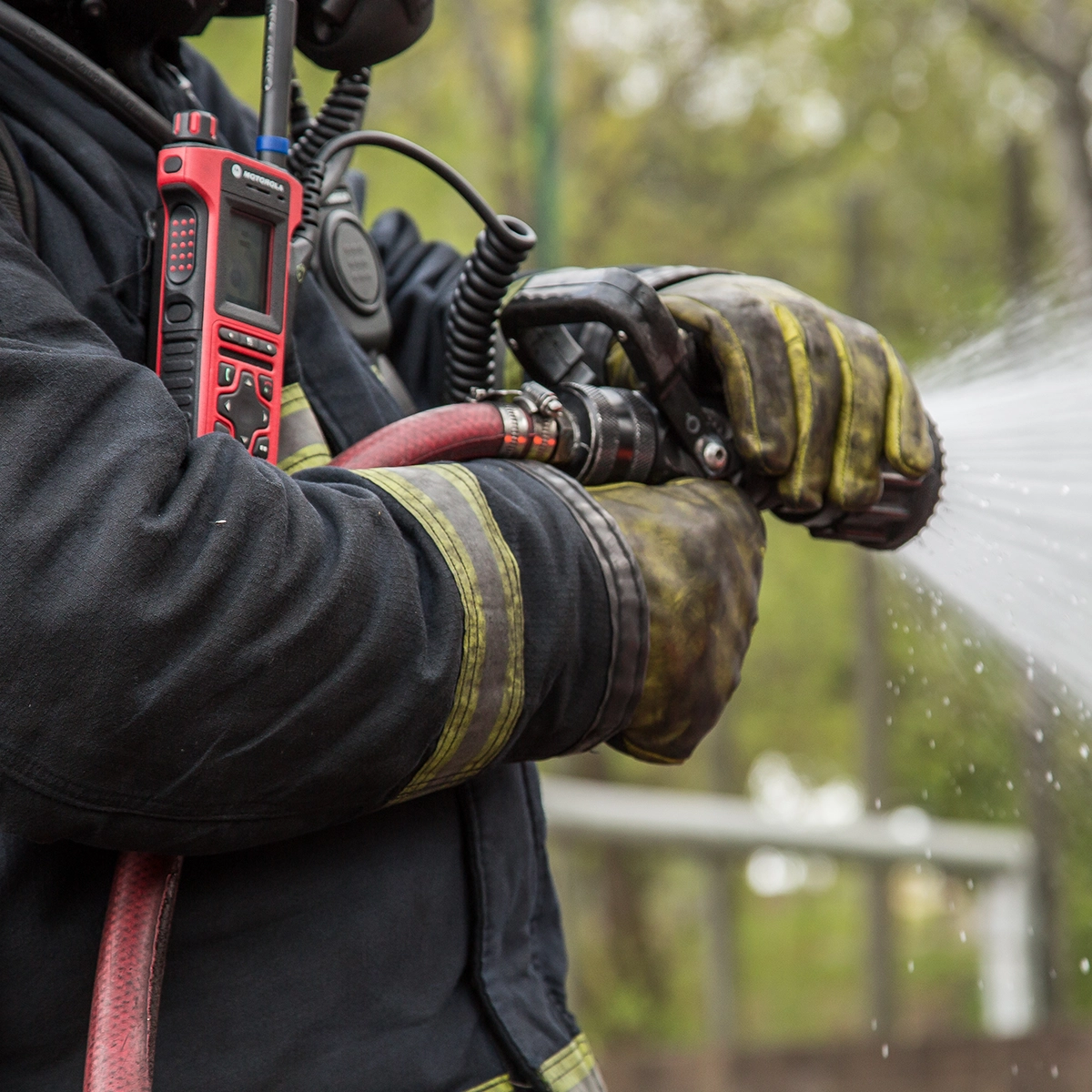 Firefighter using a hose with a red TETRA two-way radio attached to their chest.