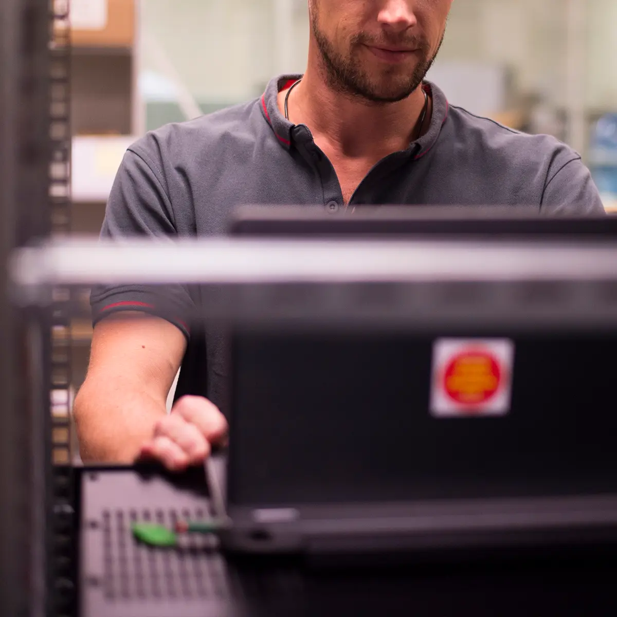 Person in a workspace wearing a dark grey CSE polo shirt with red accents, working on a laptop. Shelves and equipment are visible in the background.