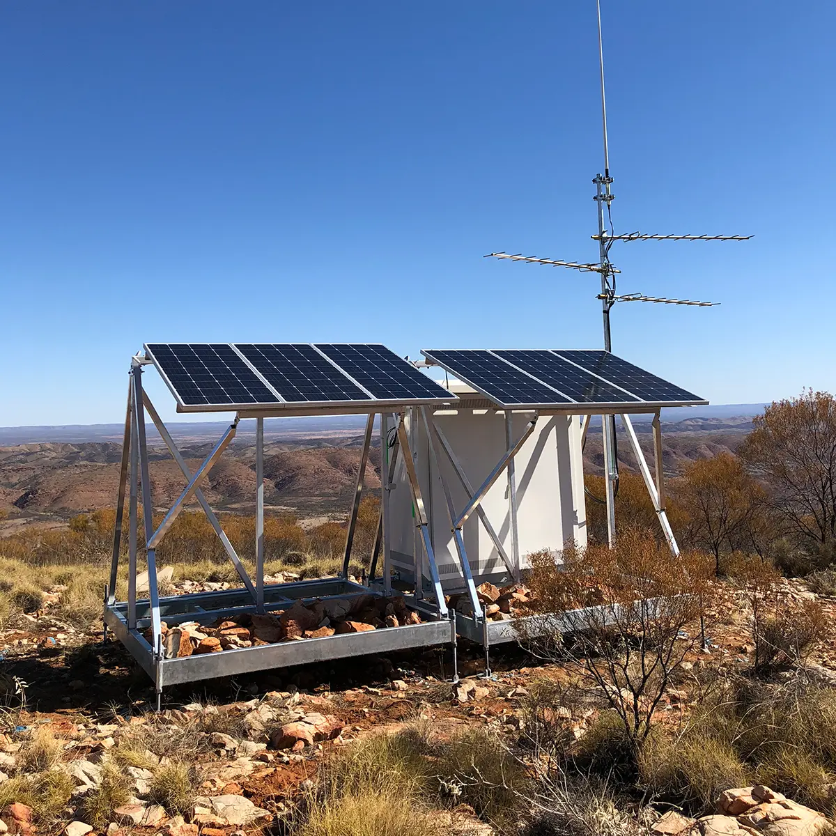 Remote installation with solar panels and an antenna in a rocky, arid landscape under a clear blue sky.