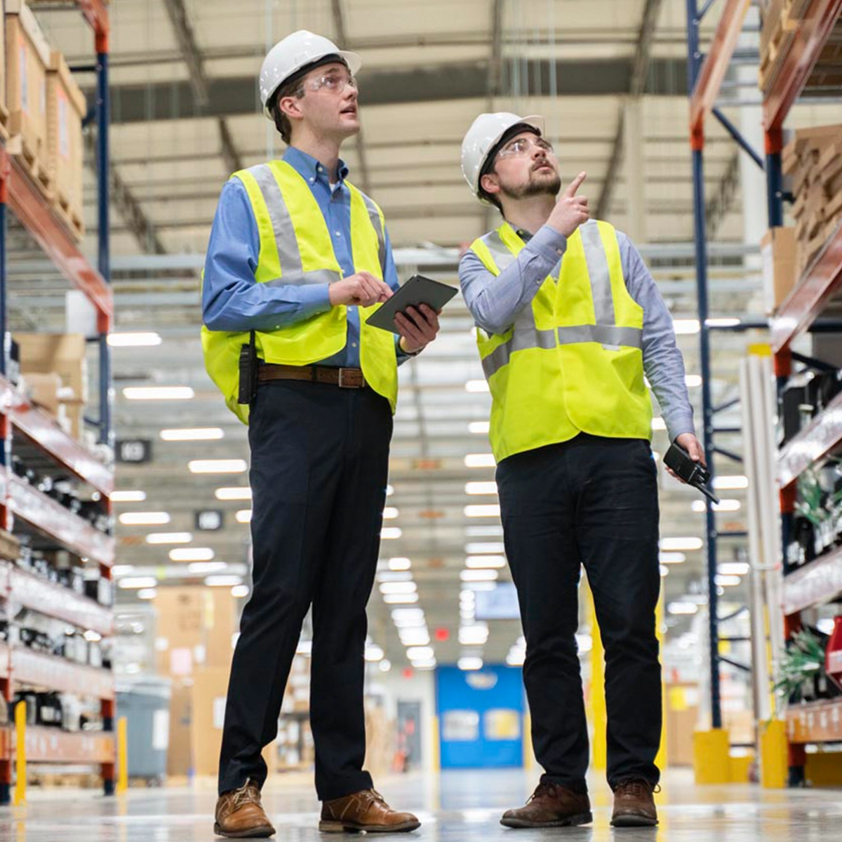 Two workers in high-vis vests and hard hats in a warehouse, one holding a tablet and the other pointing ahead.
