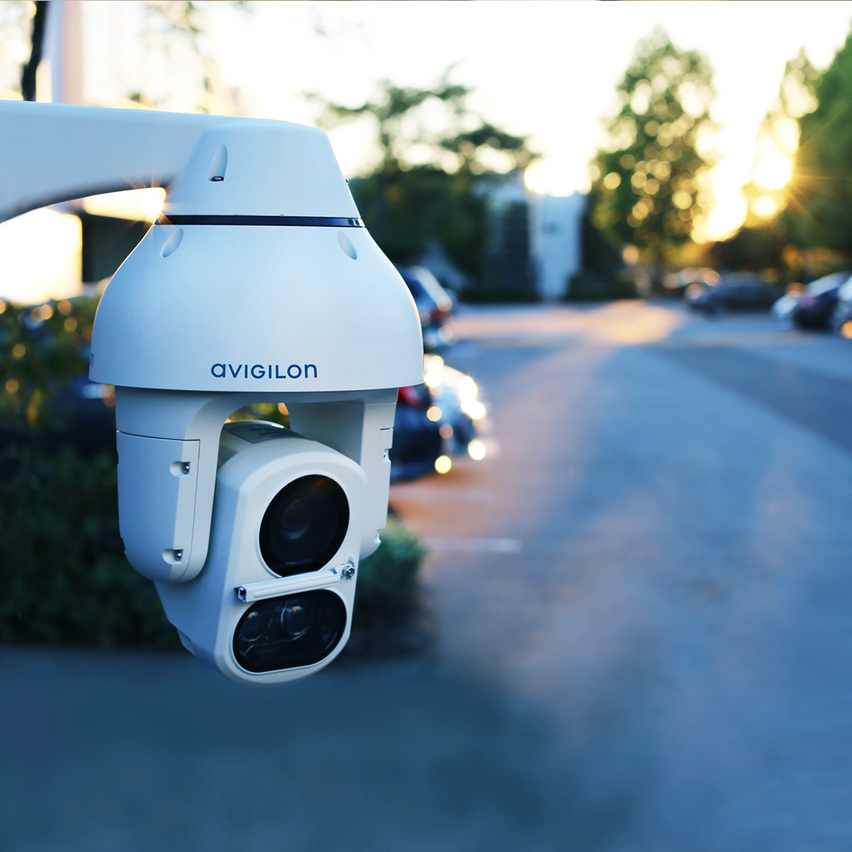 Avigilon H5A IR PTZ surveillance camera mounted outdoors with trees and a parking lot in the background.