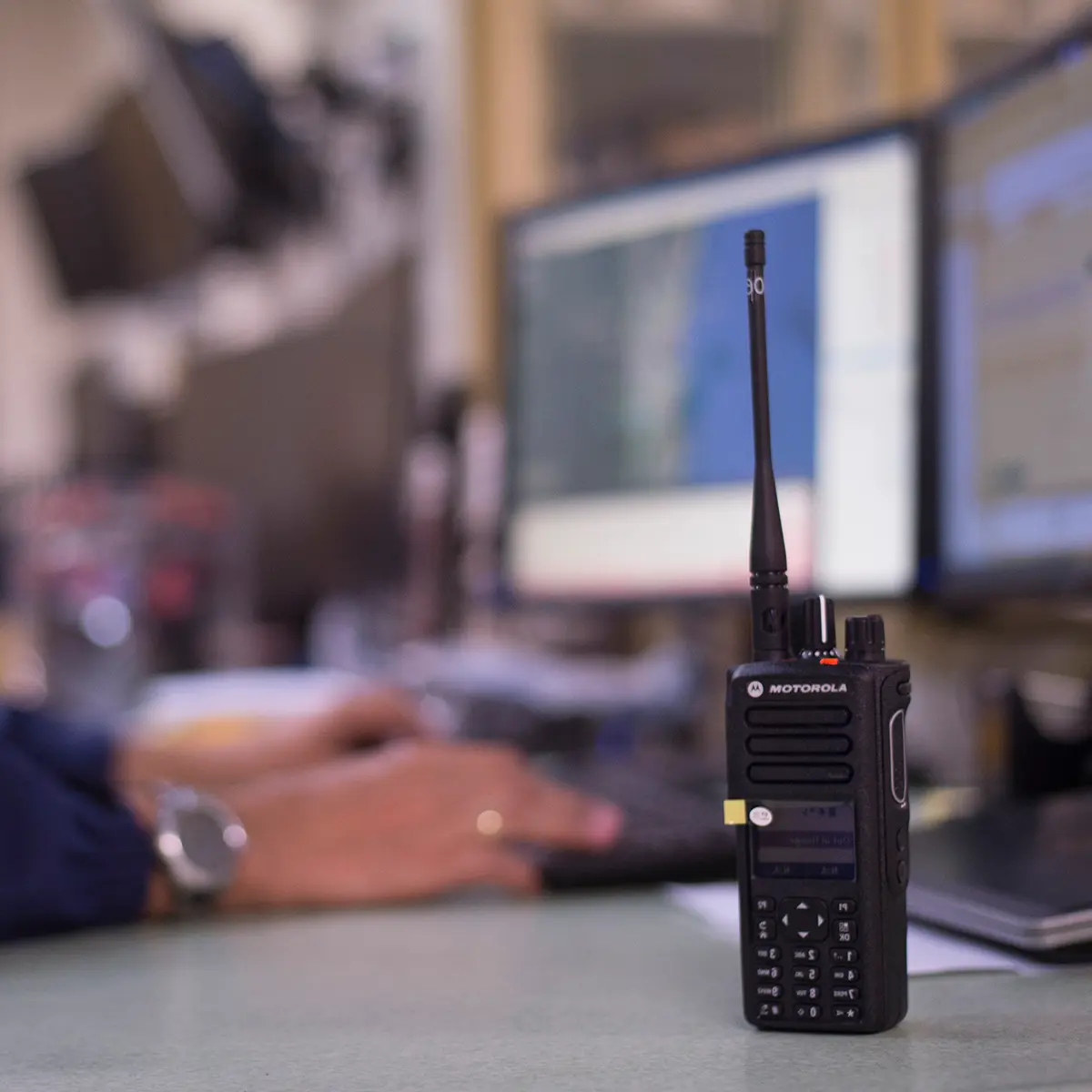 A Motorola two-way radio placed on a desk with a blurred background of computer monitors and a person working.