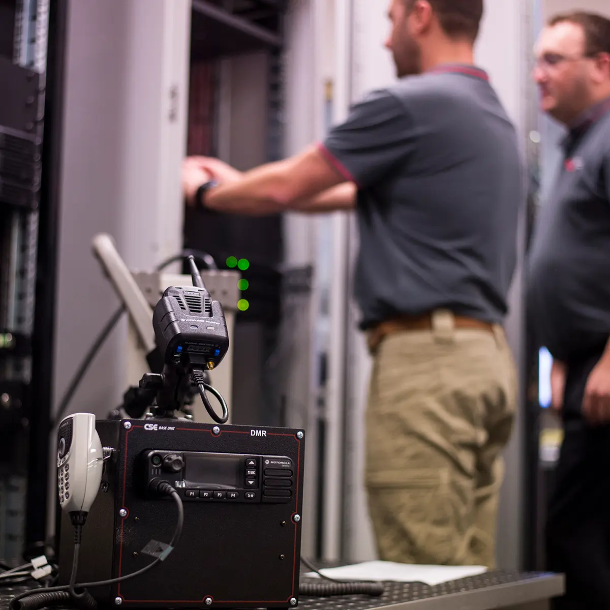 Two CSE Crosscom technicians working on server racks in a data centre, with communication equipment in the foreground.
