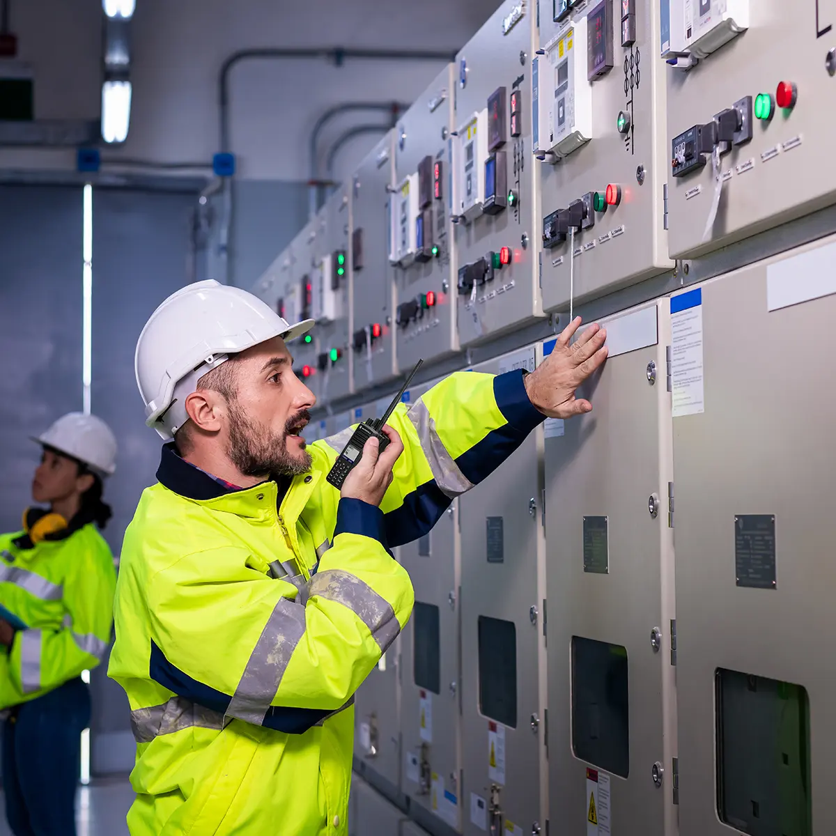 Technicians using a handheld radio and inspecting equipment in a control room, wearing safety gear.