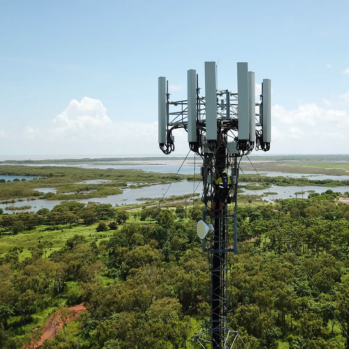 Technician in safety gear working on a telecommunications tower in a remote, green landscape with water bodies in the background. The tower features multiple large antennas.