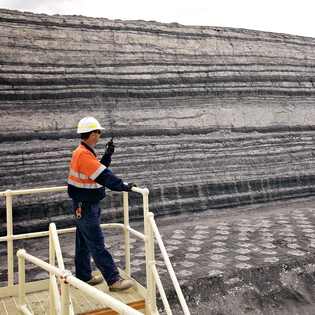 Worker in high-vis gear and hard hat using a radio on a platform overlooking a layered rock face.