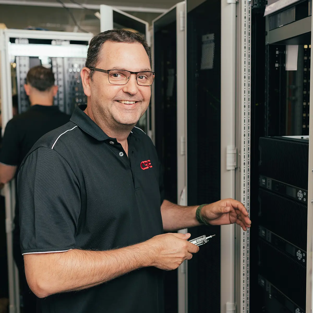 Technician working on server racks in a data centre, performing maintenance or installation. Another person is visible in the background.