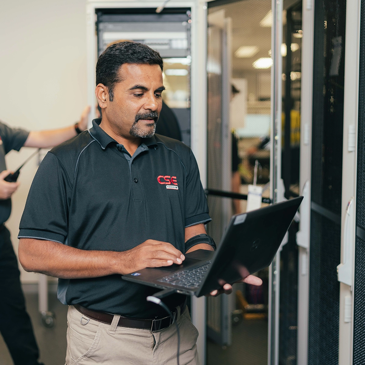 An engineer using a laptop to configure systems in a data centre, wearing CSE Crosscom branded clothing.