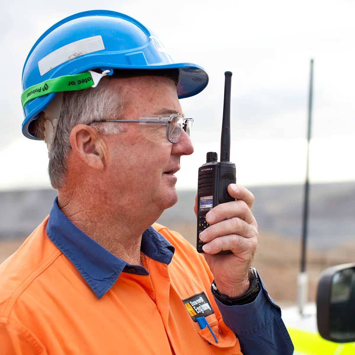 Worker in high-visibility gear and hard hat using a two-way radio for site communication.