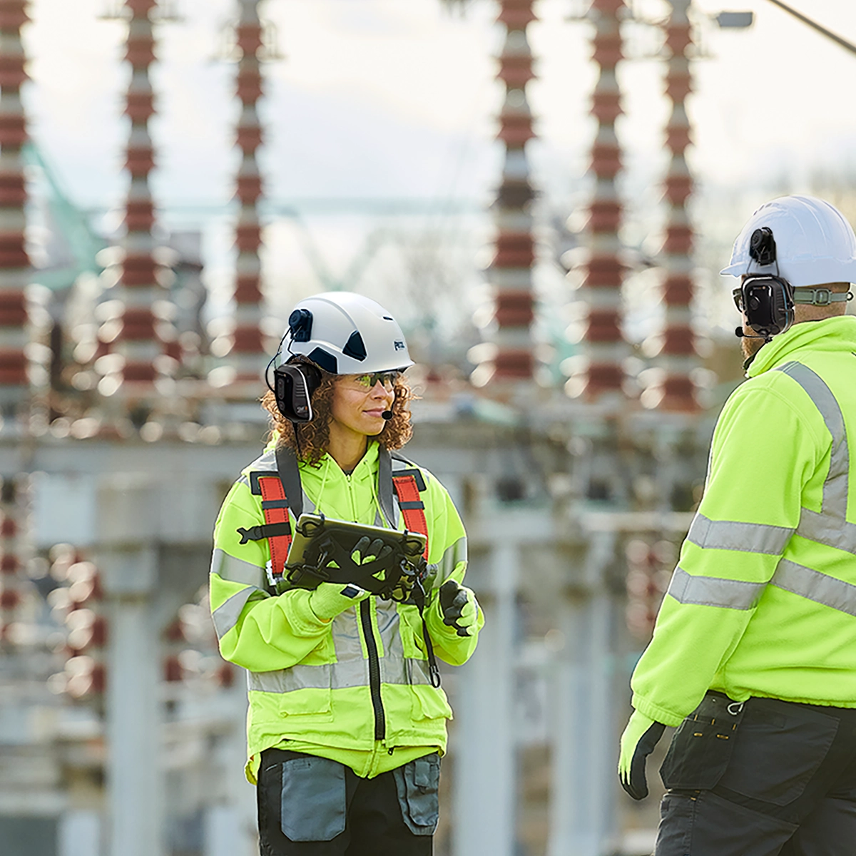 Industrial workers wearing Sensear hearing protection headsets and safety gear, communicating in a noisy worksite.