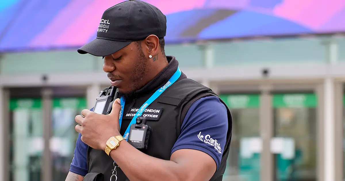 A security officer stands outside a venue, wearing a black cap, blue shirt, and a black tactical vest labeled “EXCEL LONDON SECURITY OFFICER.” Two body-worn cameras are visibly attached to the vest, with the officer holding one in their left hand. The background includes glass doors and vibrant signage, suggesting a busy, high-traffic environment. The image highlights the role of wearable technology in enhancing frontline safety and situational awareness.