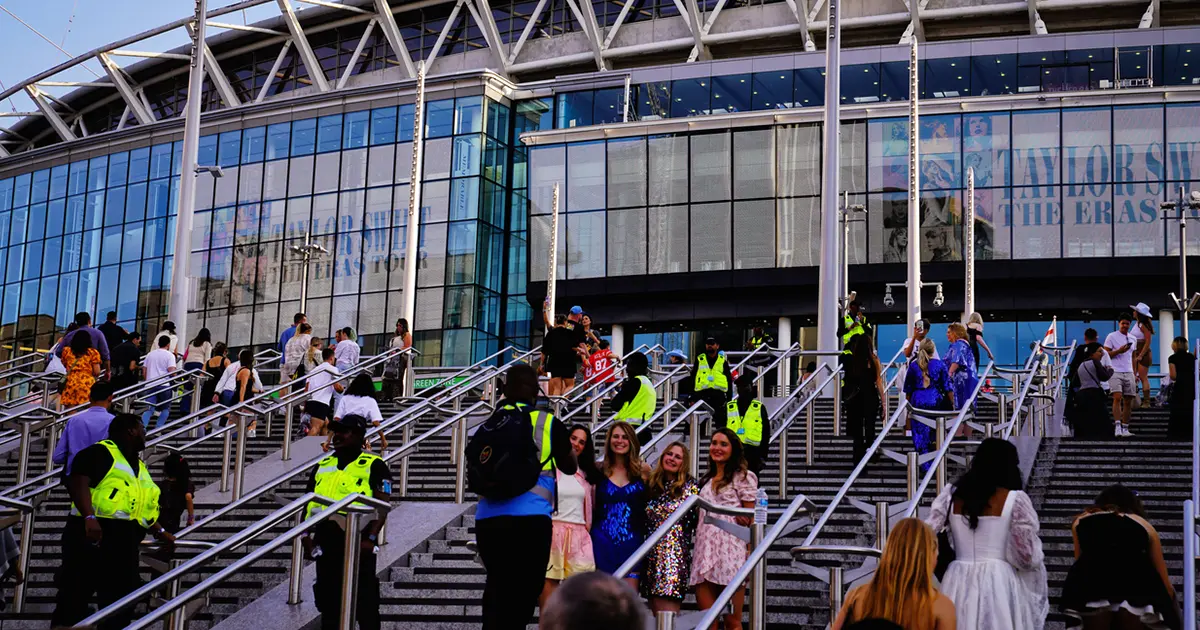 Crowd of people walking up and down stairs outside a large modern venue with glass facade. Security personnel in bright yellow jackets are present, ensuring safety. A banner for 'Taylor Swift The Eras Tour' is displayed above the entrance.