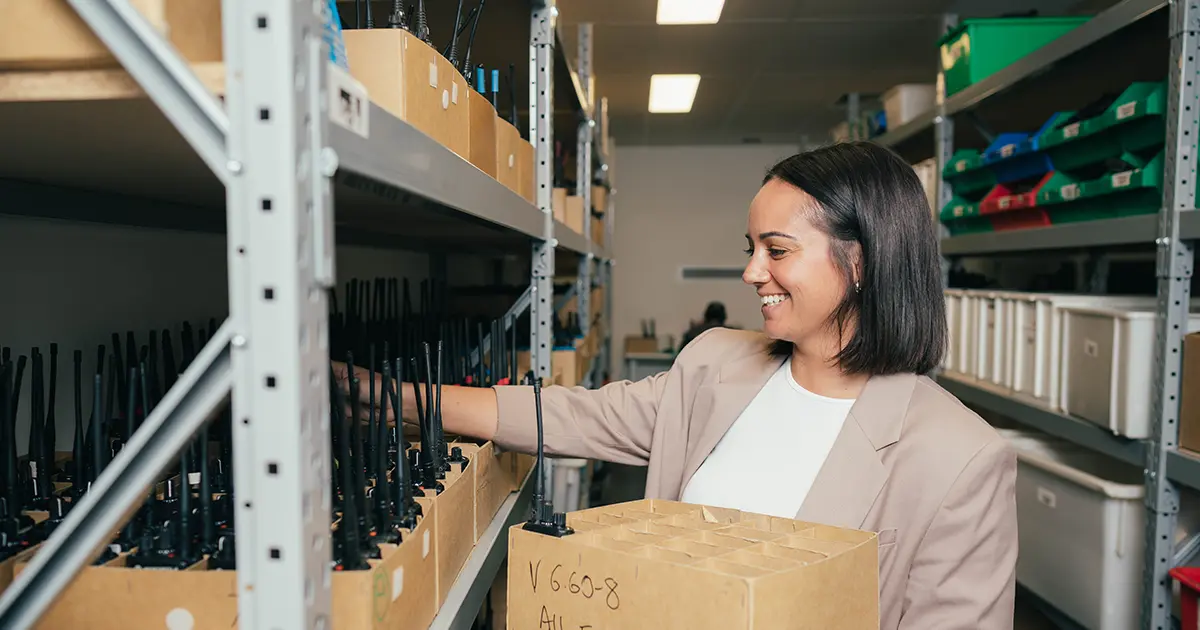 Person in a storage room reaching for a two-way radio from shelves filled with boxed units. The shelves are stocked with radios and equipment ready for deployment.