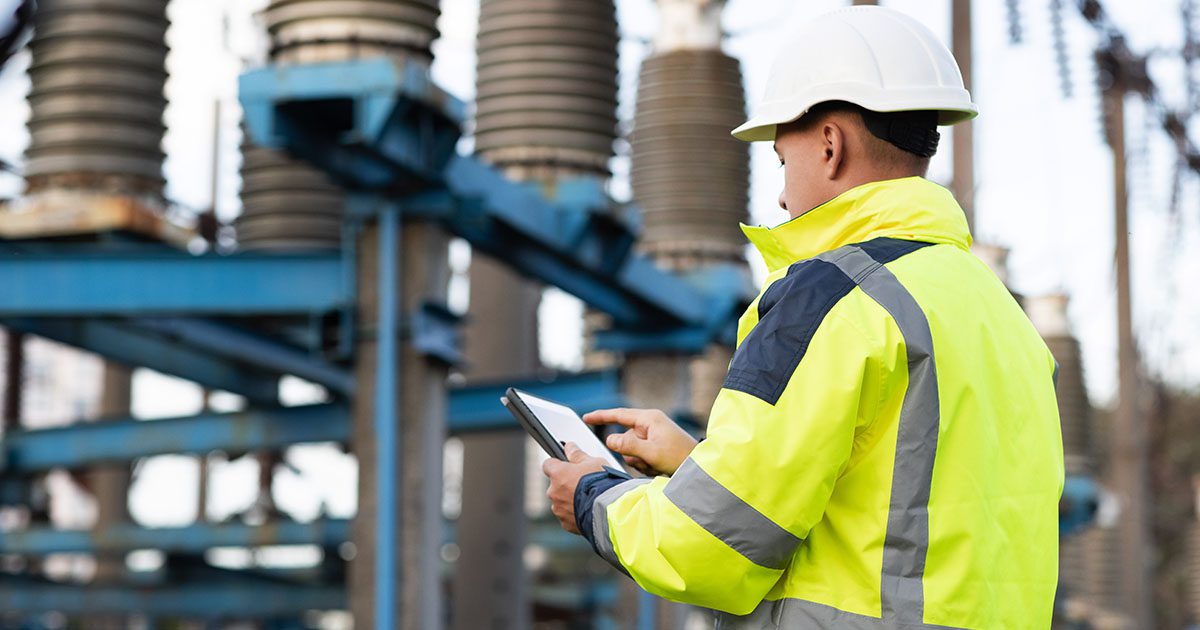 Lone utilities worker in full PPE checks the diagnostics of a remote substation on an tablet.