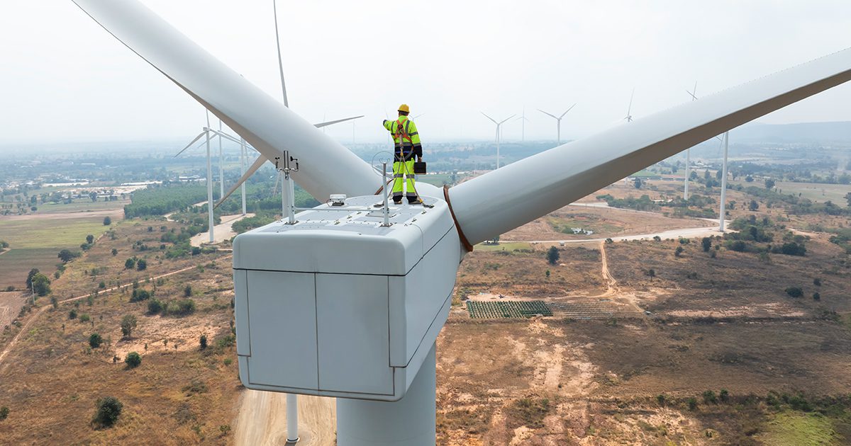 Utilities worker in full PPE is harnessed to the top of a wind turbine, completing maintenance on the machinery.