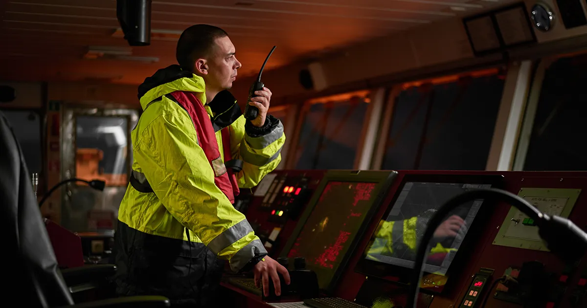 Ship captain speaks into a two way radio at the helm while consulting the navigational instruments of their large freight ship pulling into port.