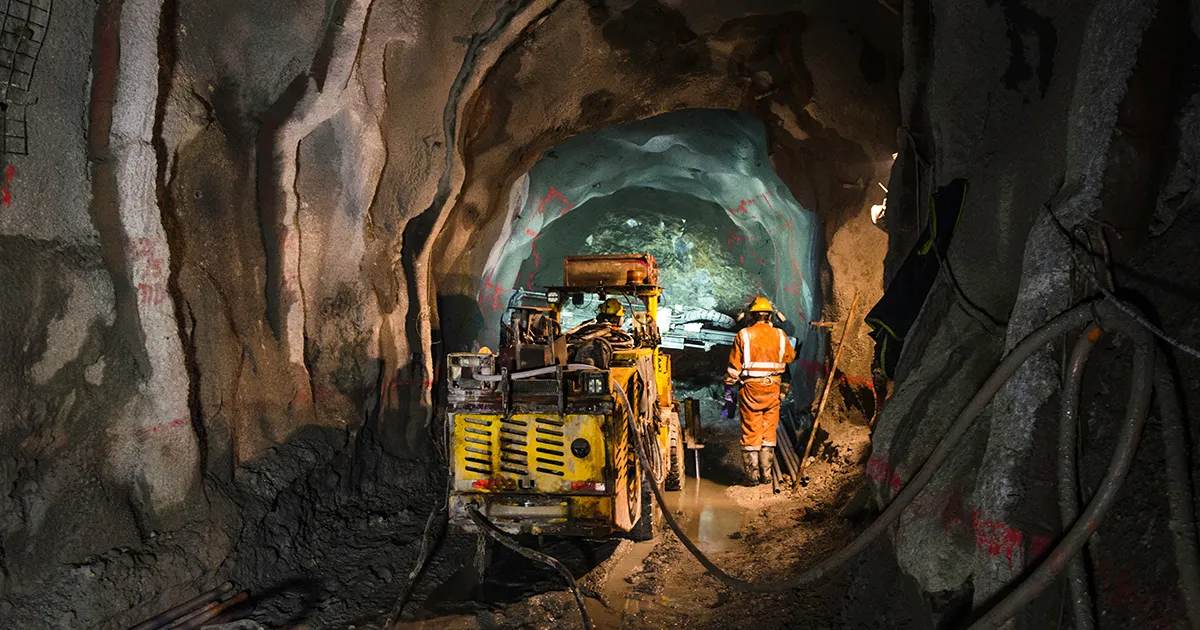 Two underground miners in High-Vis stand in a lit tunnel around mining machinery.
