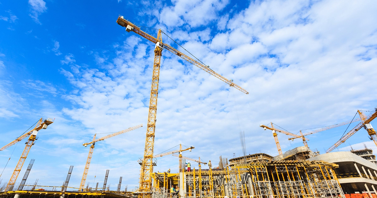Several tower cranes in operation above a busy construction site during a bright, sunny day.
