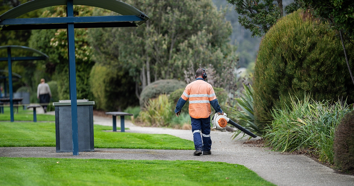 Local Council worker in high-vis clothing uses a leaf blower to clean up a local park.